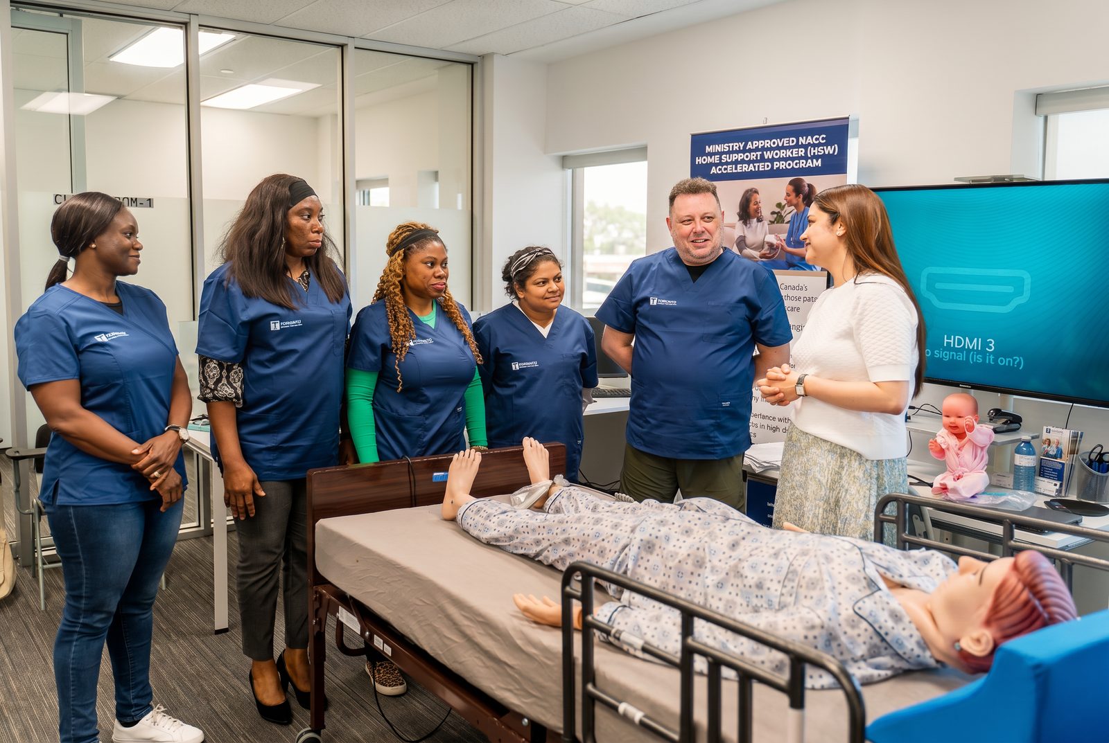 PSW students and instructor gathered around a hospital bed with a training mannequin in a bright clinical classroom